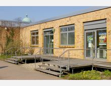 Flacher, moderner Backsteinbau mit großen Fenstern und einer Rampe für barrierefreien Zugang. Im Hintergrund ist die Kuppel eines historischen Gebäudes sichtbar, umgeben von einer gepflasterten Wegefläche und vereinzelten Pflanzen.