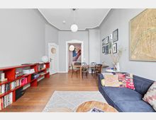 Wohnzimmer mit grauen Wänden, Holzboden und hohen Decken. Im Vordergrund steht ein dunkelblaues Sofa mit gemusterten Kissen, im Hintergrund ein Esstisch mit vier Holzstühlen; an der Wand hängen Bilder und eine große Stadtkarte, daneben ein roges Bücherregal.