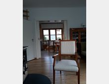 Wohnzimmer mit Holzboden, im Vordergrund ein Holzstuhl mit gestreiftem Polster. Im Hintergrund ein Durchgang zu einem Esszimmer mit Holztisch und Stühlen sowie großen Fenstern. Links steht ein Ofen, rechts ein Holzschrank.