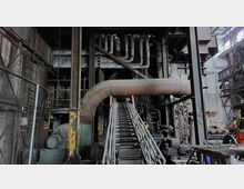 Industrielle Produktionshalle mit großer metallener Anlage, bestehend aus Rohrleitungen, Stahlträgern und einer schrägen Metalltreppe in der Mitte. Im Hintergrund sind Wände aus Ziegeln und Beton sowie zahlreiche technische Bauteile sichtbar.