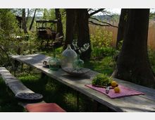 Holztisch mit rustikaler Bank in einem schattigen Waldgarten, dekoriert mit Glasvasen, frischen Blumen, Kräutern und Obst. Im Hintergrund sind Bäume, Sträucher und eine alte Holzkutsche sichtbar.