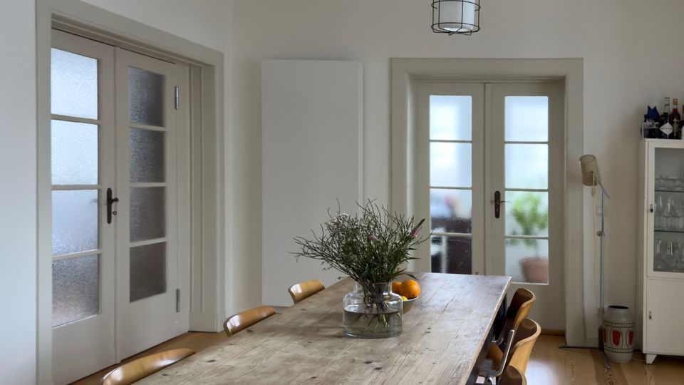 Heller Essbereich mit langem Holztisch und mehreren Holzstühlen auf hellem Dielenboden. Zwei Glastüren mit Sprossen, eine hängende Deckenlampe und ein weißer Geschirrschrank prägen den Raum.