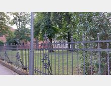 Metallzaun mit dekorativen Verzierungen umgibt eine Grünfläche mit Bäumen, im Hintergrund ein rotes Backsteingebäude und ein helles Gebäude.