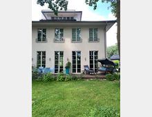 Moderne Hausfassade mit weißen Wänden und großen bodentiefen Fenstern. Vor dem Haus befindet sich ein Garten mit Rasen, einer Holzterrasse, Sitzmöbeln und Pflanzen.