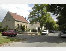 Straßenszene in einem Wohngebiet mit freistehenden Einfamilienhäusern, schrägdächern und beigen Fassaden. Die Straße ist von Bäumen gesäumt, mit geparkten Autos auf beiden Seiten, und ein kleiner gepflasterter Platz mit Steinen und parkendem roten Auto ist im Vordergrund sichtbar.