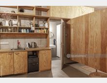 Kleine Küche mit offenen Holzregalen, in denen Tassen, Gläser und Lebensmittel aufbewahrt werden. Darunter befinden sich eine Arbeitsplatte mit Spüle, Wasserkocher und Schränken; rechts führt eine Tür zu einem kleinen Badezimmer mit Waschbecken und Spiegel.