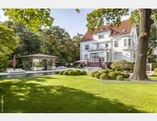 Außenansicht einer eleganten Villa mit weißer Fassade und rotem Dach, umgeben von einer gepflegten Gartenlandschaft mit einem großen Rasen, geformten Buchsbaumsträuchern, einem modernen Pavillon mit Glaswänden und einem langen Pool. Im Hintergrund sind Bäume und Sonnenschirme zu sehen, die eine entspannte Atmosphäre schaffen.