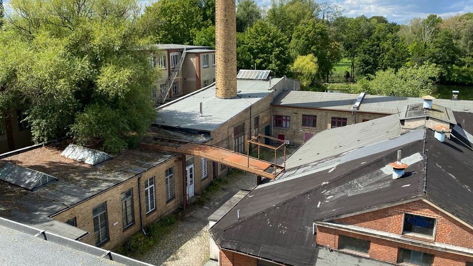 Blick von oben auf ein ehemaliges Industrieareal mit flachen Backsteingebäuden, dunklen Dachflächen und einem hohen gemauerten Schornstein; eine erhöhte Metallbrücke verbindet zwei Gebäudeteile über einem gepflasterten Hof. Im Hintergrund liegen dichte Bäume und Grünflächen unter einem teils bewölkten Himmel.