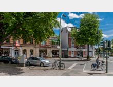 Straßenszene mit historischen zweigeschossigen Backsteinhäusern, Geschäften im Erdgeschoss und Baumreihen am Straßenrand. Im Vordergrund eine Kreuzung mit Ampeln, einem parkenden Auto, Fahrrädern sowie einer Person auf einem Fahrrad.