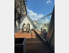Eine moderne Dachterrasse mit Holzboden, flankiert von Glasgeländern, mehreren dekorativen Skulpturen und Pflanzen in Pflanzgefäßen. Im Hintergrund ist ein blauer Himmel mit wenigen Wolken sichtbar, und städtische Gebäude sind entlang des Horizonts erkennbar.