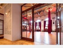 Ein heller Festsaal mit hohen Rundbogenfenstern, roten Vorhängen und Holzboden, gesehen durch geöffnete Glastüren. Die Raumdecke ist mit sichtbaren Stahlträgern und Beleuchtungselementen gestaltet.