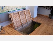 Ein Raum mit Holzdielenboden und einer geöffneten Holzklappe, die zu einem Schacht mit gemauerten Wänden führt. An der Wand hängt eine große Karte, daneben sind Kleidungsstücke aufgehängt.