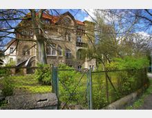 Alt-Text: Historisches Wohnhaus mit sandfarbenem Mauerwerk, roten Ziegeldächern und Rundbogenfenstern, umgeben von einem Garten mit Bäumen und Sträuchern. Ein Metallzaun grenzt das Grundstück zur Straße hin ab.