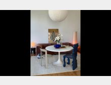 Modernes Wohnzimmer mit einem runden weißen Tisch, auf dem eine Vase mit weißen Lilien steht. Im Hintergrund befinden sich ein braunes Sofa, eine minimalistische Kunstwerk-Leinwand an der Wand und eine großformatige Papierlampe an der Decke.