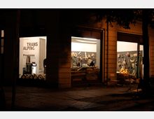 Außenansicht eines Schaufensters bei Nacht mit Einblick in eine beleuchtete Galerie oder Ausstellung. Innen sind Kunstwerke und Objekte aus Holz und anderen Materialien an Wänden und Tischen sichtbar, die mit dem Schriftzug "Transalpino Open Progress" beworben werden.