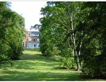 Ein großes, rotes Backsteingebäude mit großen Fenstern und einem gewölbten Eingang im Erdgeschoss steht am Ende einer weitläufigen, grasbewachsenen Fläche, umgeben von hohen Bäumen und dichten Sträuchern. Die Szene befindet sich in einem parkähnlichen, grünen Gelände.