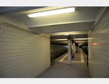 Ein U-Bahnsteig mit beigen Kachelwänden, einer abgehängten Decke mit Leuchtstofflampen und einer Bahnsteigkante, die durch weiße Fliesenstreifen markiert ist. Ein Stahlpfeiler mit Graffiti und Beschilderung ist zentral sichtbar, im Hintergrund eine leicht gebogene Tunnelröhre.