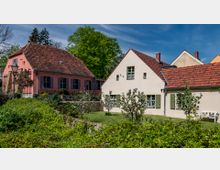 Zwei kleine Häuser mit roten Ziegeldächern in einem ländlichen Garten mit grünen Bäumen und Sträuchern. Das linke Haus ist rosa mit blauen Fensterläden, das rechte beige mit grünen Fensterläden.
