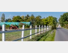 Ländliche Szene mit einer Wiese, eingefasst von einem weißen Holzzaun, einem roten Traktor und einem grünen Folientunnel im Hintergrund. Rechts verläuft eine asphaltierte Landstraße, die von Bäumen gesäumt ist; im Hintergrund steht ein Windrad.