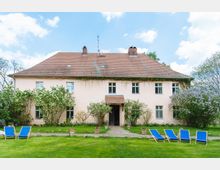 Großes, zweistöckiges Gebäude mit cremefarbener Fassade und rotem Ziegeldach, umgeben von Fliederbüschen und gepflegtem Rasen. Vor dem Haus stehen mehrere blaue Liegestühle auf der grünen Wiese.