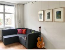 Wohnzimmer mit einer grauen Eckcouch, dekoriert mit bunten Kissen, und einer akustischen Gitarre auf einem Ständer daneben. Im Hintergrund befinden sich weiße Vorhänge vor einem großen Fenster und drei gerahmte Bilder an der Wand.