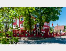 Rotes Gebäude mit verspielten architektonischen Details, umgeben von einer roten Mauer im Stil einer Burg mit Wappensymbolen. Im Vordergrund stehen Bäume, außerdem sind zwei Straßenschilder mit den Aufschriften „Seepromenade“ und „Lessingallee“ sowie ein benachbartes Gebäude mit rotem Ziegeldach sichtbar.