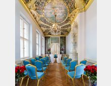 Ein prunkvoller Raum mit blau-goldenen Stühlen, einer dekorativen bemalten Decke mit Goldakzenten, einem Kronleuchter und einem holzvertäfelten Boden; im Hintergrund ein geschmückter Tisch vor einer verzierten Doppeltür und bemalten Wanddekorationen.