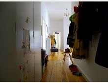 Langer, schmaler Flur mit hellem Holzfußboden und weißen Wänden. Links steht ein Schrank mit befestigten Zeichnungen, rechts hängen Jacken und Helme sowie ein gelber Stuhl, am Ende des Flurs sind eine Garderobe und eine Tür zu sehen.