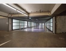 Ein moderner Parkhausbereich mit Betonboden, niedriger Decke und heller Deckenbeleuchtung. Die Wände sind teilweise aus mattem Glas mit Metallrahmen, und ein langer schmaler Gang führt im Hintergrund weiter.