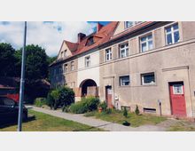 Zweistöckiges Wohngebäude mit rotem Satteldach und beige-brauner Fassade, mehreren Fenstern und einer roten Eingangstür. Vor dem Haus befinden sich ein kleiner Garten mit Büschen und eine gepflasterte Gehwegfläche, im Hintergrund stehen Bäume.