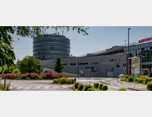 Rundverkehr vor einem modernen runden Bürogebäude mit Glasfassade; im Vordergrund ein Kreisverkehr mit Blumenbeeten und Straßenmarkierungen. Rechts ist ein Bahnhofsgebäude mit einem „Bahnhof“-Schriftzug und Parkhinweisen sichtbar.
