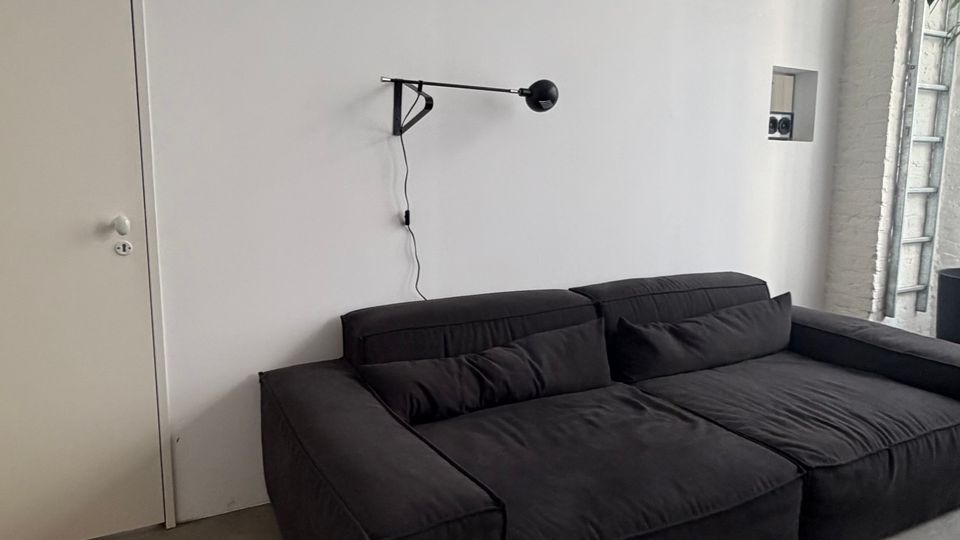 Minimalistisches Loft-Wohnzimmer mit weißer Trennwand und hohen Oberlichtern, Betonboden und hohen Decken. Vor der Wand steht ein großes schwarzes Sofa; rechts ist eine Backsteinwand mit hohen Fenstern und eine angelehnte Leiter zu sehen.