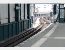 Bahnsteig einer erhöhten S-Bahn-Station mit dem Schild "Gleisdreieck", Sicht auf gebogene Schienen und umliegende Stahlkonstruktionen. Im Hintergrund sind moderne Gebäude und Teile der Bahnsteigüberdachung erkennbar.