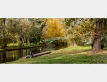 Ein Park mit einem kleinen Fluss, über den eine grüne Fußgängerbrücke führt. Der Fluss ist von Bäumen mit herbstlichem Laub umgeben, und im Vordergrund befindet sich eine Holzplattform am Ufer.