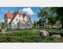 Wohnhaus mit Satteldach und hellem Putz, umgeben von einem Holzzaun und einem gepflegten Garten mit blühenden Rosen. Im Vordergrund befinden sich große Steine, einer davon mit einer Plakette, sowie ein kleiner Park mit Bäumen und Gebüsch.