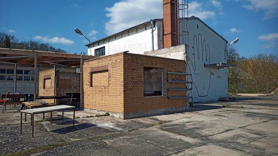 Ein industrielles Gelände mit einem flachen, gelben Backsteingebäude und einem weißen Gebäude im Hintergrund mit Wandzeichnung einer Hand. Der Betonboden ist teilweise verwittert, und mehrere freistehende Tische stehen unter einer Überdachung.