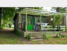 Eine kleine, einfache Gartenhütte mit grün gestrichenen Türen und Fensterläden, umgeben von Bäumen. Die Hütte hat eine überdachte Veranda mit einer Wäscheleine, einigen Möbeln und einer Treppe, die in einen grasbewachsenen Bereich führt.