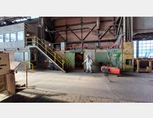 Innenansicht einer Industriehalle mit rohen Backsteinwänden, Metallgerüsten und großen Fenstern rechts. Im Vordergrund ein Schutzanzug-Modell, eine rostige Metallwand und ein roter Druckbehälter, links führt eine Treppe mit gelbem Handlauf nach oben.