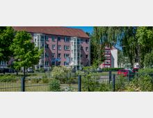 Mehrstöckiges Wohngebäude mit rosa Fassade und rotem Satteldach, mit verglasten Treppenhausaufgängen und mehreren Fenstern. Davor liegen eine Grünfläche mit Bäumen und Sträuchern sowie ein Zaun und geparkte Autos; im Hintergrund ist ein weiteres Gebäude mit roten Rahmen zu sehen.