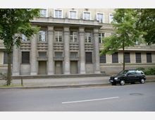 Außenansicht eines historischen Gebäudes mit einer symmetrischen Fassade aus Stein, bestehend aus massiven Säulen und dekorativen Elementen. Vor dem Eingang befinden sich Stufen, und ein schwarzes Auto ist am Straßenrand geparkt; Bäume säumen die Straße.