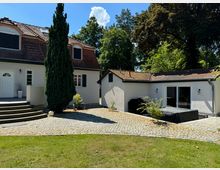 Weißes Einfamilienhaus mit roten Ziegeldächern und mehreren Dachfenstern, umgeben von einem gepflasterten Hof und grünem Rasen. Im Vordergrund eine Treppe zum Eingang und rechts eine Terrasse mit Gartenmöbeln, im Hintergrund hohe Bäume.
