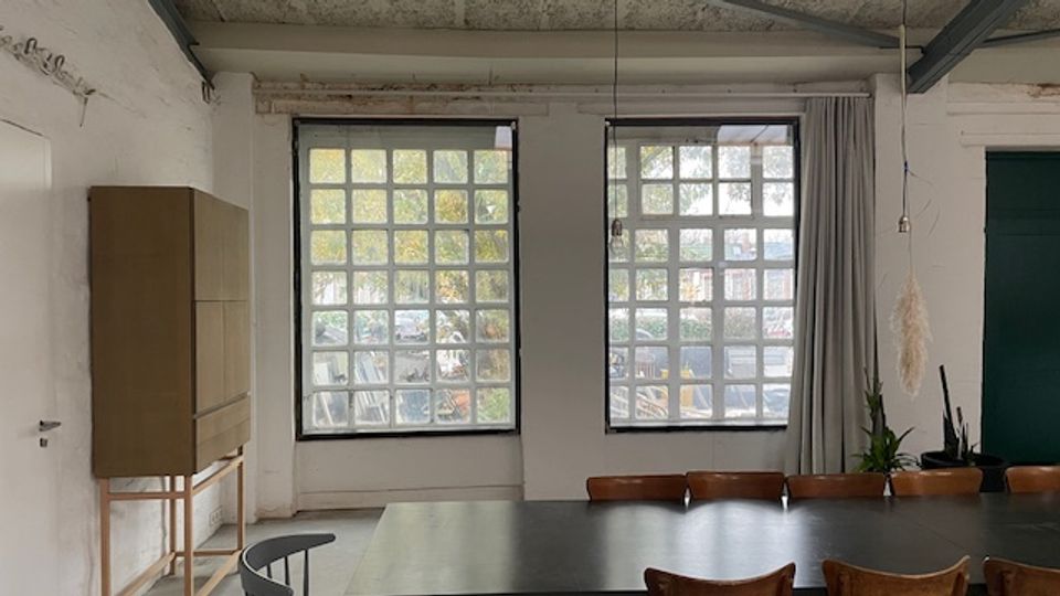 Schlichter Innenraum mit zwei großen, gitterförmigen Fensterfronten, die Tageslicht hereinlassen. Vor den Fenstern steht ein langer Konferenztisch mit Stühlen; links ein hoher Schrank und rechts Vorhänge sowie einige Pflanzen vor einer dunklen Tür.