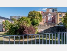 Ein Stadtbild mit einer Grünfläche im Vordergrund, umgeben von Gebäuden. Im Hintergrund sind ein rosafarbenes Gebäude mit weißen Details und weitere Wohnhäuser zu sehen, teils mit modernem und teils mit älterem Baustil.