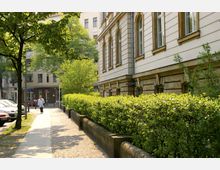 Straßenszene in einer städtischen Umgebung mit einem historischen Gebäude auf der rechten Seite, das durch verzierte Fassadendetails und hohe Fenster geprägt ist. Links ein gepflasterter Gehweg mit Bäumen und geparkten Autos, daneben eine gepflegte Hecke.