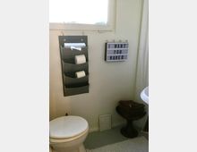 Ein schlichtes Badezimmer mit weiß gestrichenen Wänden, einer Toilettenschüssel im Vordergrund und einem kleinen grauen Wandorganizer für Toilettenpapier. Rechts daneben hängt ein dekoratives Schild mit dem Text "I WANT YOU NAKED", darunter steht ein Hocker mit gefalteten Handtüchern. Oben befindet sich ein Fenster, das Tageslicht hereinlässt.