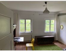 Ein schlicht eingerichteter Raum mit Holzboden und weiß gestrichenen Wänden. Zwei Fenster mit weißen Vorhängen lassen Tageslicht in den Raum, der mit einem schwarzen Tisch, einem gelben Stuhl, einer antiken Holztruhe und einem kleinen runden Spiegel ausgestattet ist.