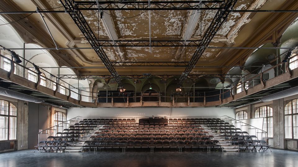 Großer, leerer Veranstaltungssaal mit einer zentralen Tribüne aus gestaffelten Sitzreihen und offenem Bühnenboden im Vordergrund. Hohe Decke mit abblätterndem Putz, sichtbarer Traversenkonstruktion und umlaufenden Galerien sowie großen Rundbogenfenstern entlang der Seitenwände.