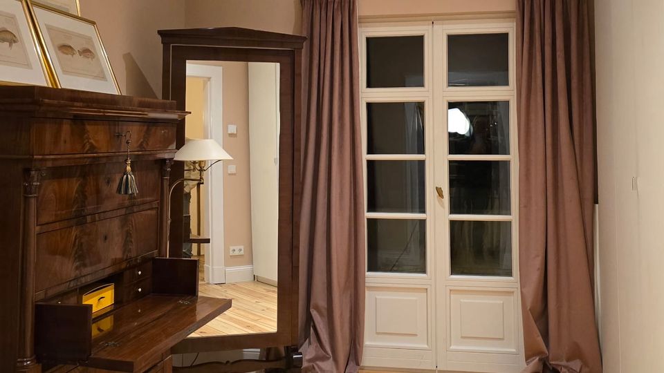 Kleines Zimmer mit hellem Holzdielenboden, beigefarbenen Wänden und einer weißen doppelflügeligen Balkontür mit braunen Vorhängen. Links stehen ein dunkler Holzsekretär und ein Spiegel-/Schrankmöbel, rechts eine schlichte weiße Wand.