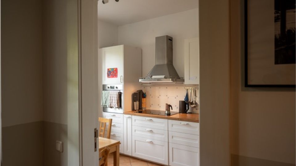 Blick durch eine offene Tür in eine helle Küche mit weißen Schränken, Holzarbeitsplatte und Edelstahl-Dunstabzugshaube über dem Herd. Fliesenboden, kleine Küchenutensilien auf der Arbeitsfläche und ein Holztisch mit Stühlen sind sichtbar.