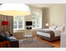 Helles Schlafzimmer mit einer modernen Einrichtung, bestehend aus einem Doppelbett, einem Sofa, einem kleinen Couchtisch und einer Wandhalterung mit Flachbildfernseher. Große Fenster mit Vorhängen lassen Tageslicht herein; der Boden ist mit hellem Holz und einem Teppich versehen.